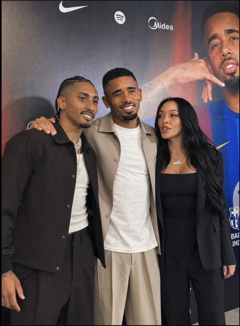 A man resembling Gabriel Jesus posing at a promotional event beside two other people, with brand logos visible behind them.