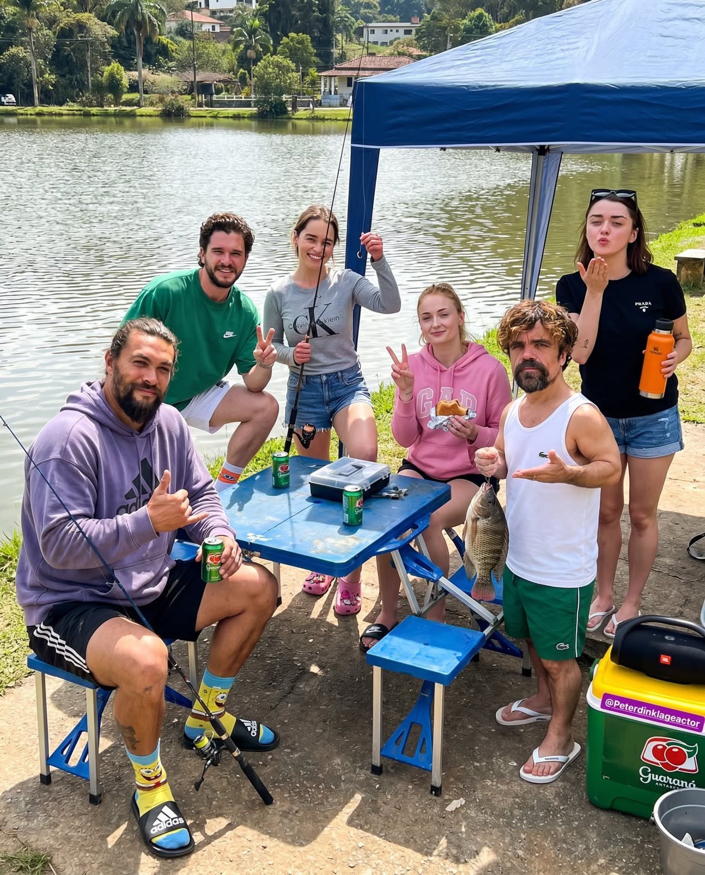 Five people resembling Jason Momoa, Peter Dinklage, Kit Harington, Sophie Turner, and Emilia Clarke posed together at a lakeside picnic.