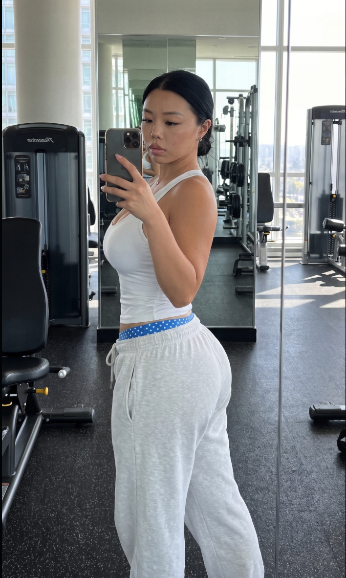 A woman in a white tank top and sweatpants taking a mirror selfie in a gym.
