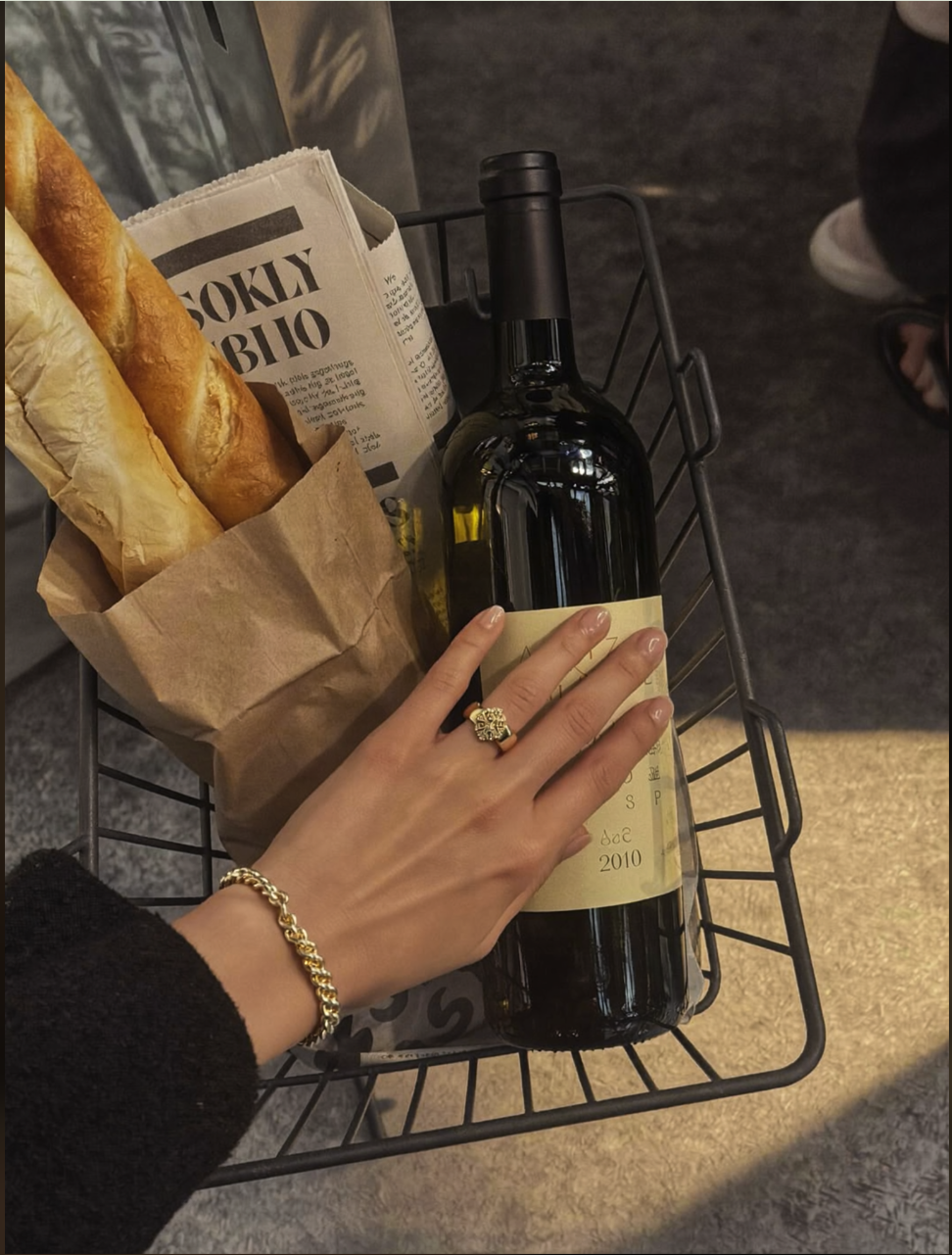 A manicured hand with a gold ring and bracelet resting on a wine bottle in a black wire basket holding a wrapped baguette.