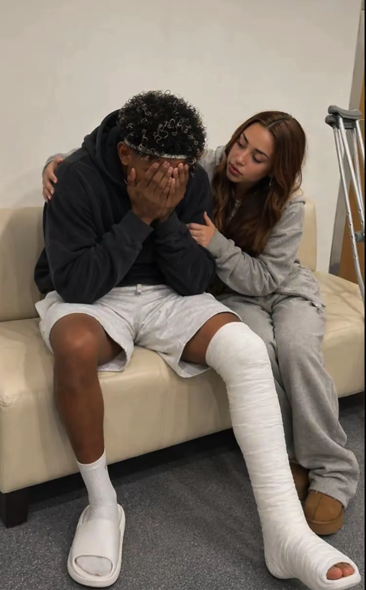 Lamine Yamal with a bandaged leg seated on a couch with his face in his hands, comforted by a woman beside him.