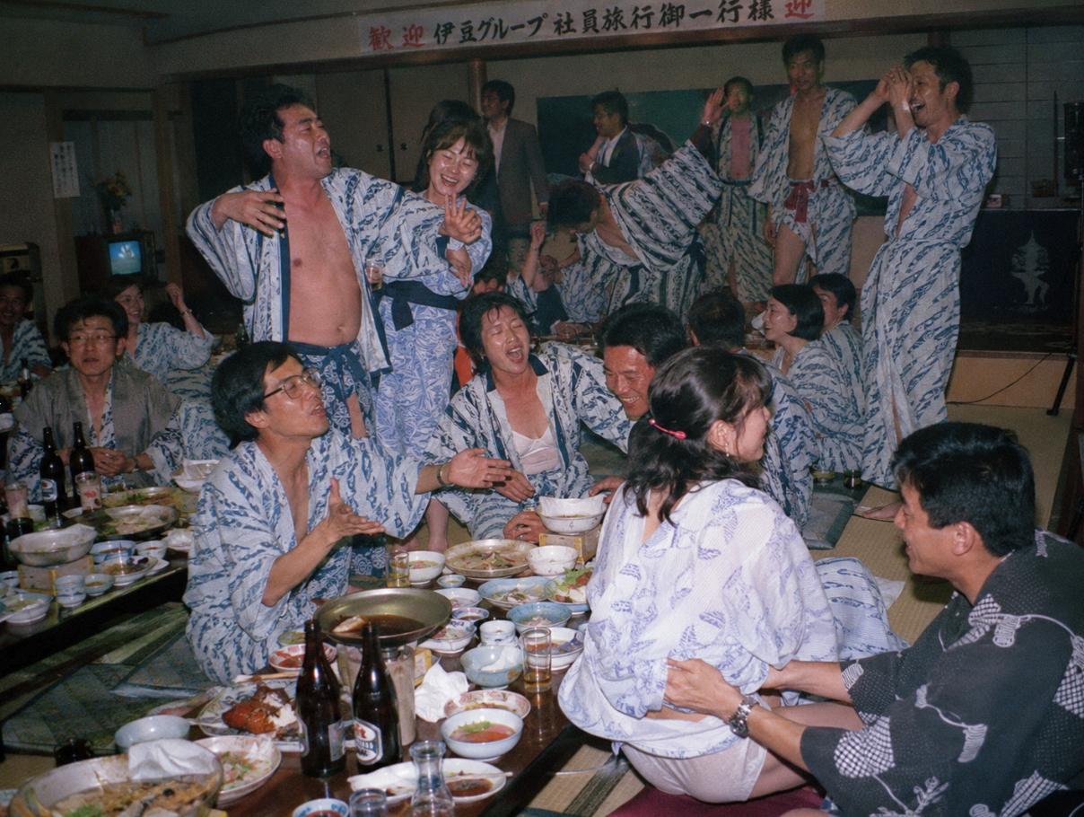 A group of people resembling Hiroshi Itsuki, Jun Kunimura, and Claude Maki in matching yukata at a banquet table with a Japanese welcome banner overhead.
