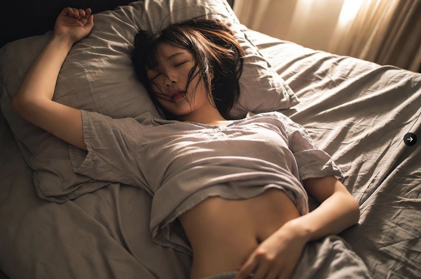 A woman asleep on a bed, with warm light through curtains.