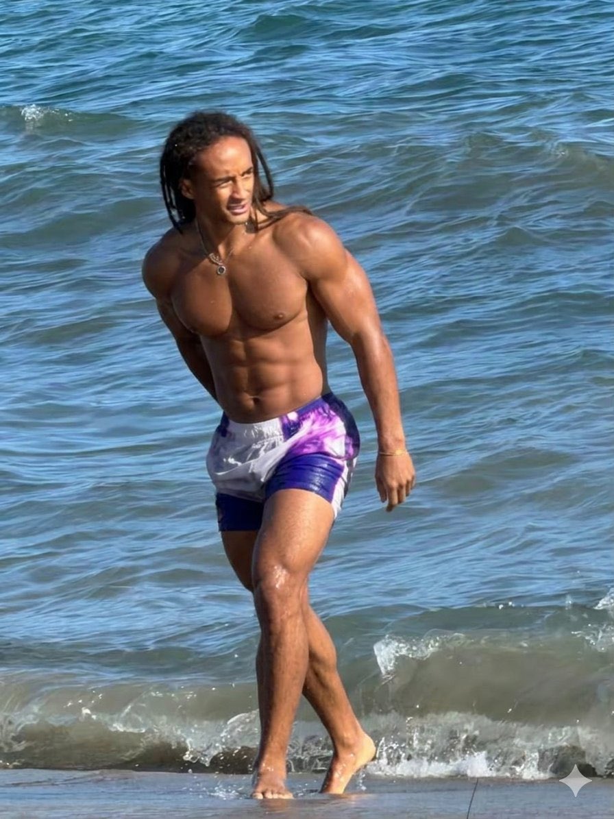 A shirtless man resembling Jaden Smith standing at the water's edge on a beach.