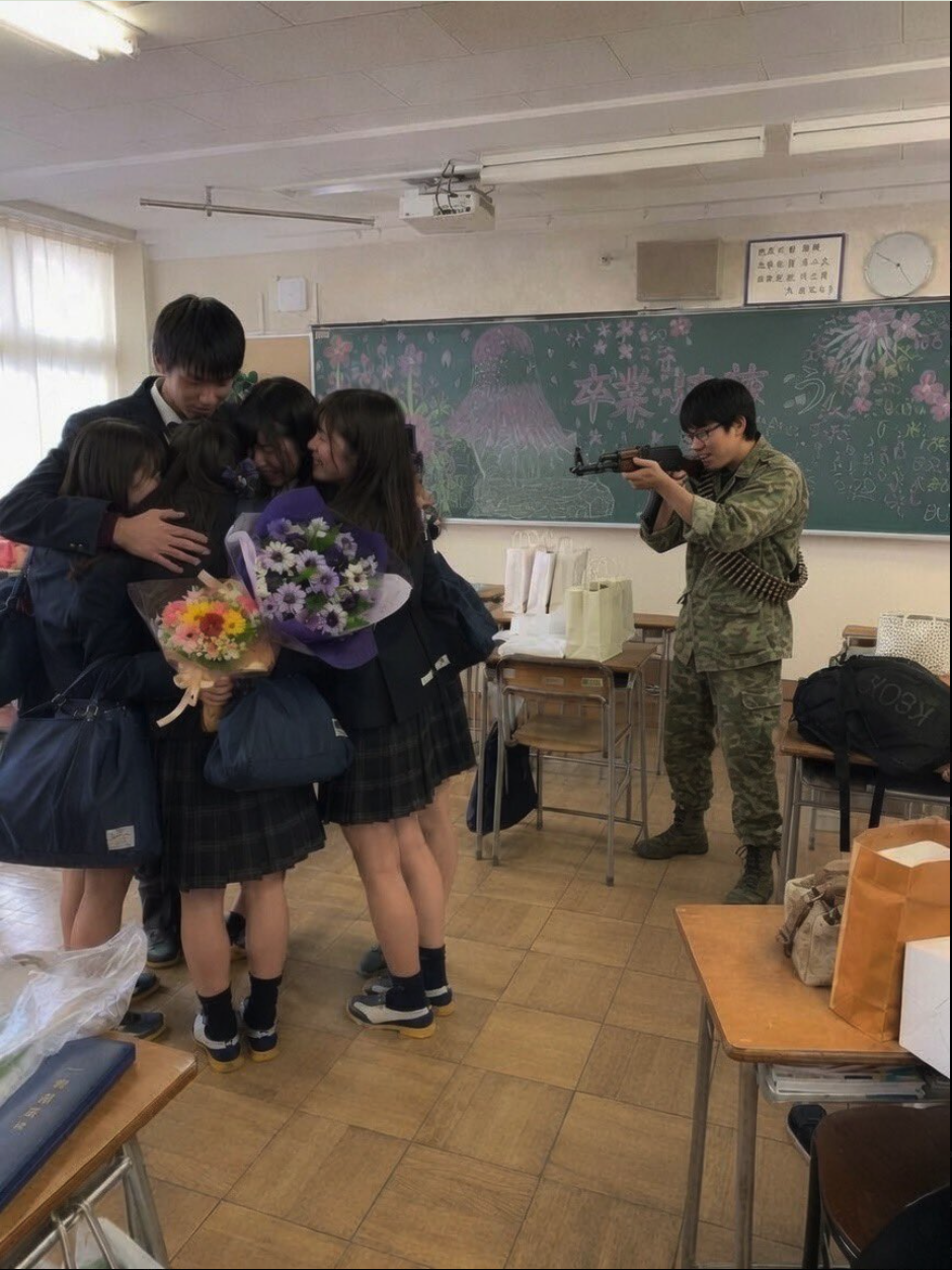 Students in dark Japanese school uniforms holding flower bouquets huddle together in a classroom while a person in camouflage clothing aims a rifle at them.