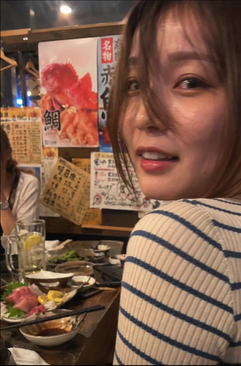 A young woman in a striped shirt turned toward the camera inside a Japanese restaurant with seafood menu posters behind her.