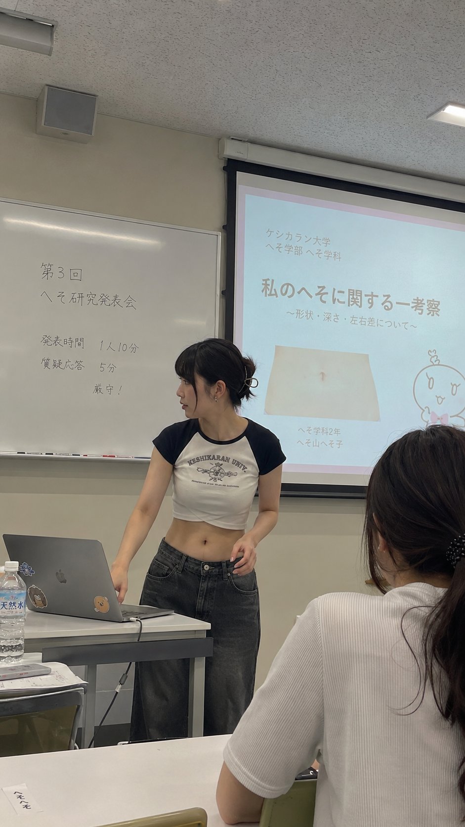 A young woman in a crop top standing at a desk in a classroom, presenting beside a projection screen.