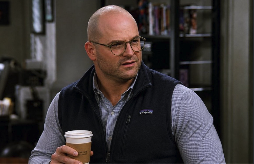 A man resembling Jason Alexander, wearing glasses and a black vest, holding a paper coffee cup indoors.