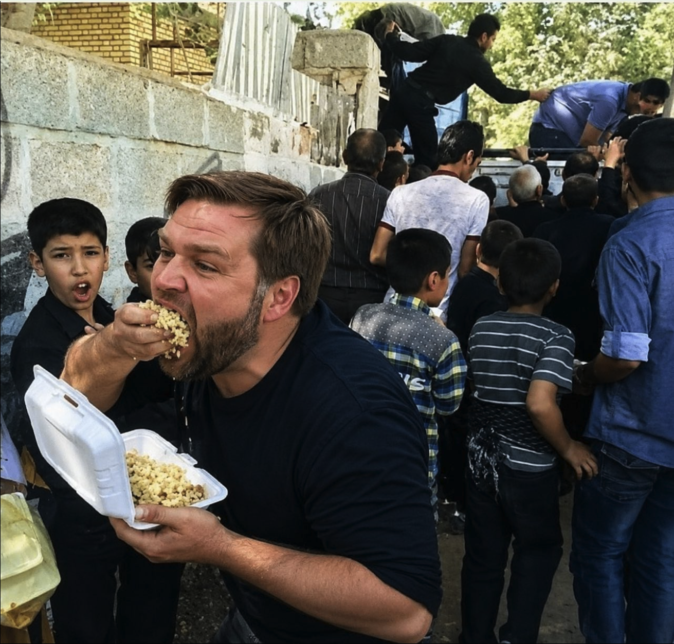 A man resembling JD Vance eating grain from a styrofoam takeout container amid a crowd of people outdoors.