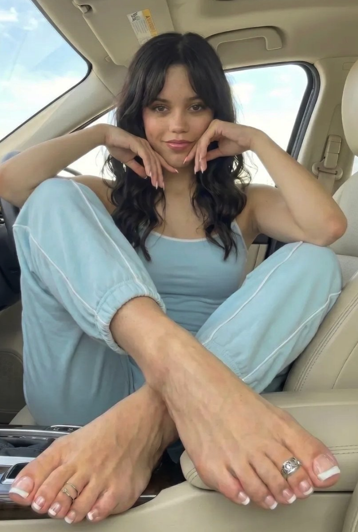 A woman resembling Jenna Ortega seated inside a car, resting her hands under her chin.