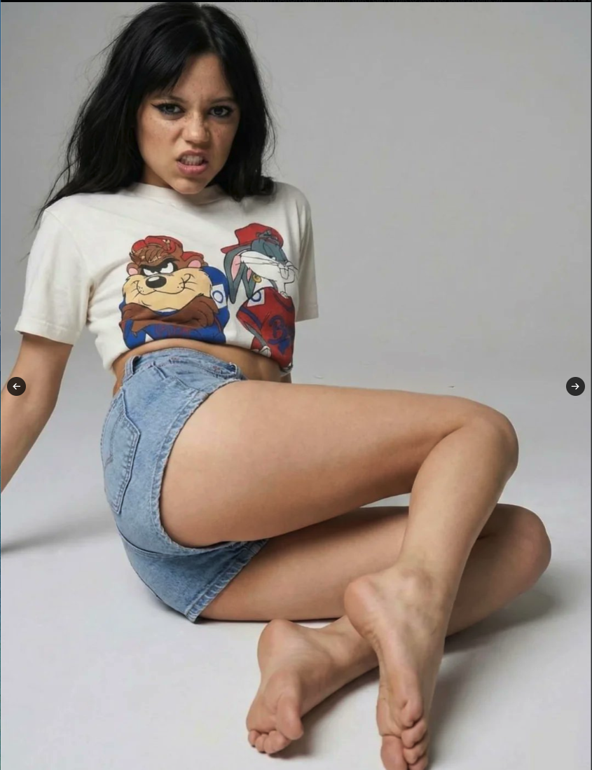 A woman resembling Jenna Ortega seated on a studio floor in a Looney Tunes crop top and denim shorts.
