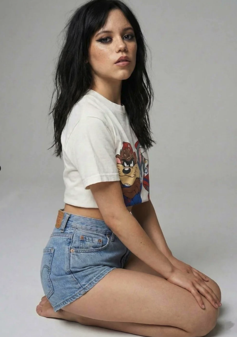 A woman resembling Jenna Ortega kneeling against a plain backdrop, wearing a cropped white Tasmanian Devil T-shirt and denim shorts.