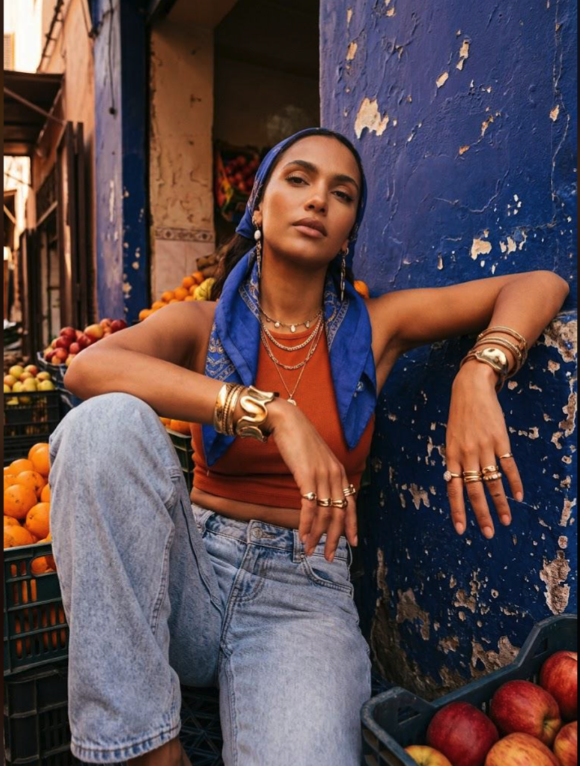 A woman seated against a weathered blue wall beside crates of fruit.