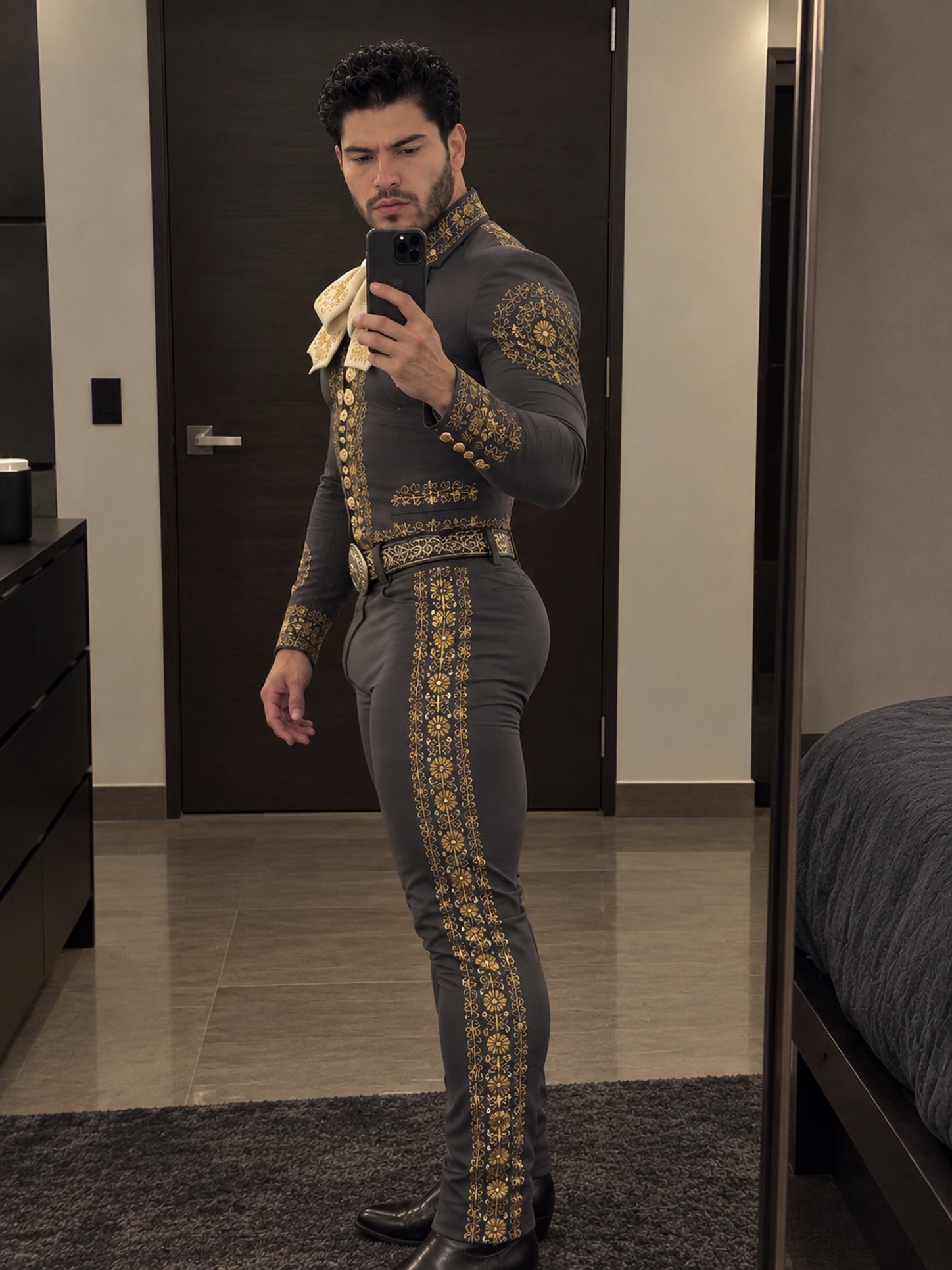 A man resembling Jesús Alonzo taking a mirror selfie while wearing an ornate gray and gold charro suit.