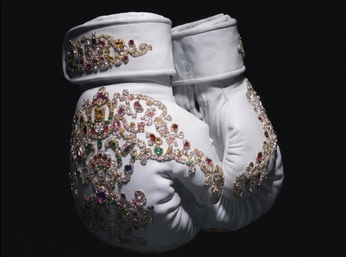 A pair of white boxing gloves decorated with elaborate gold and gemstone embellishments, displayed against a black background.