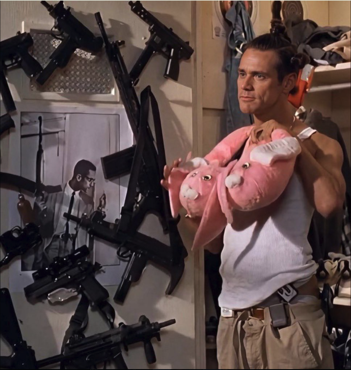 A man resembling Jim Carrey holding two pink stuffed animal toys in front of a wall covered with firearms.