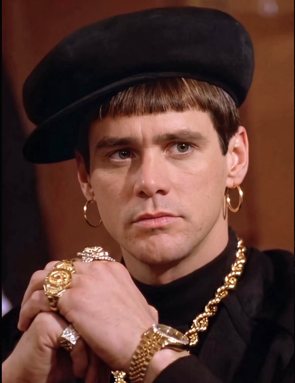 A man resembling Jim Carrey wearing a black beret, gold jewelry, and a dark top, posed with hands near his chin.
