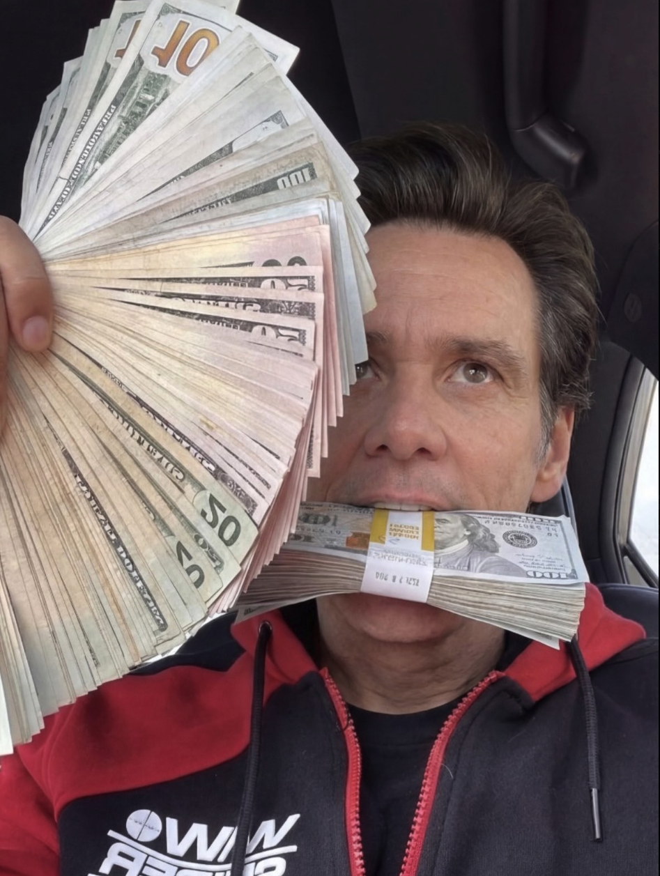 A man resembling Jim Carrey holding a fan of US dollar bills and a banded stack of $100 bills in his mouth, seated inside a car.
