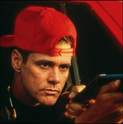 A man resembling Jim Carrey wearing a red baseball cap backward while looking down at a phone.