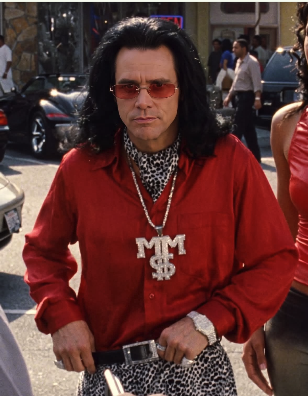 A person resembling Jim Carrey with long dark hair, red-tinted rectangular sunglasses, a red shirt, leopard-print scarf, and a large MM$ pendant necklace.