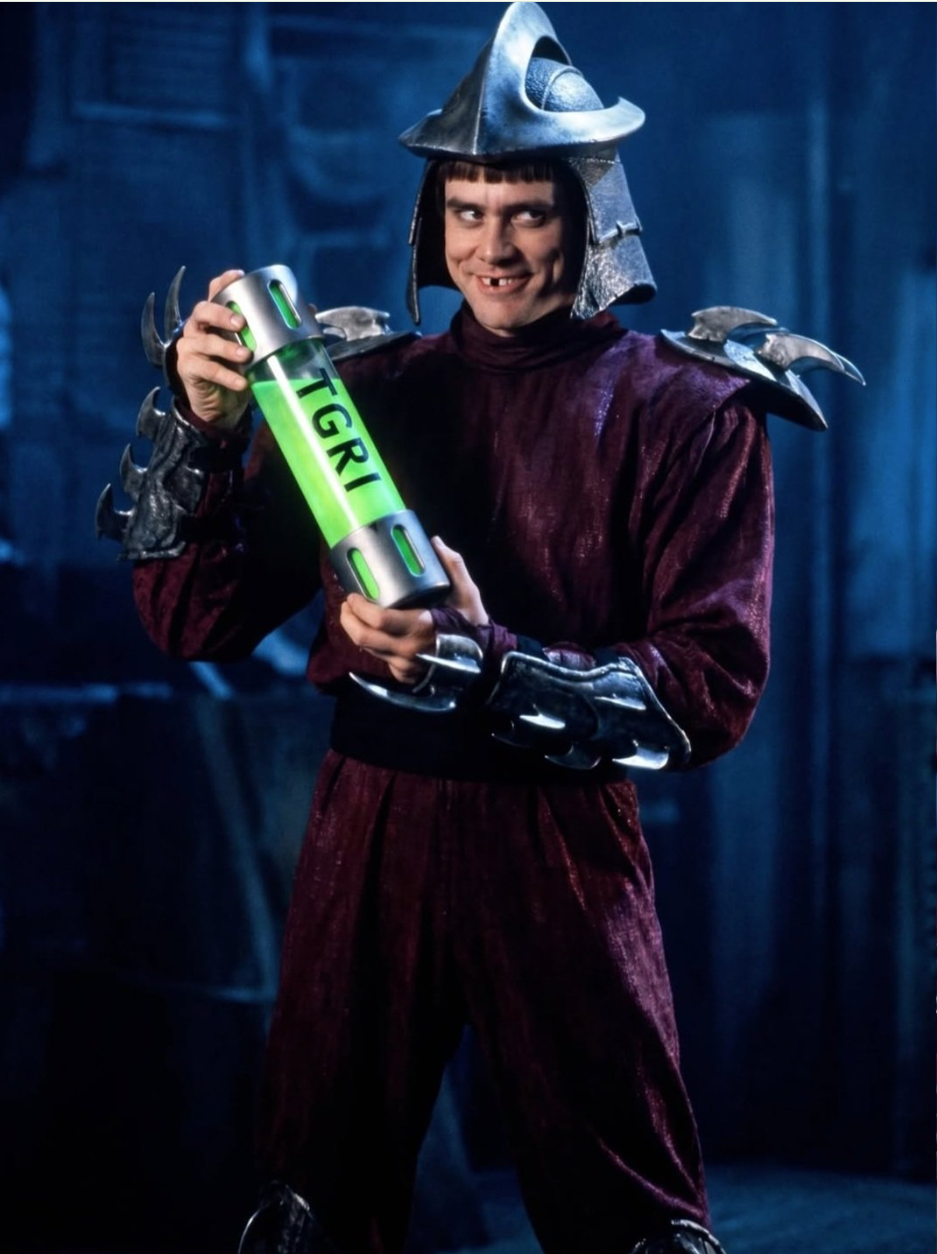 A man resembling Jim Carrey in maroon medieval-style armor and horned helmet, holding a glowing green canister labeled TGRI.