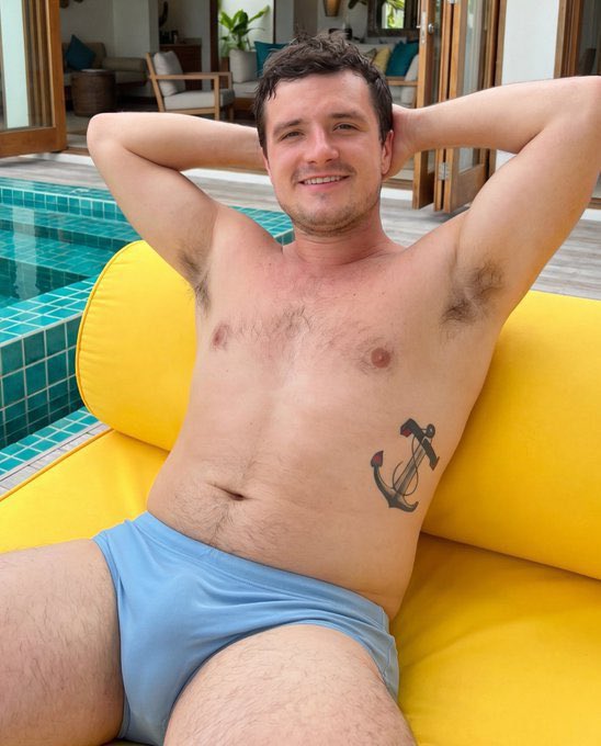 A man resembling Josh Hutcherson reclining shirtless on a yellow pool float in swim trunks by a tiled pool.