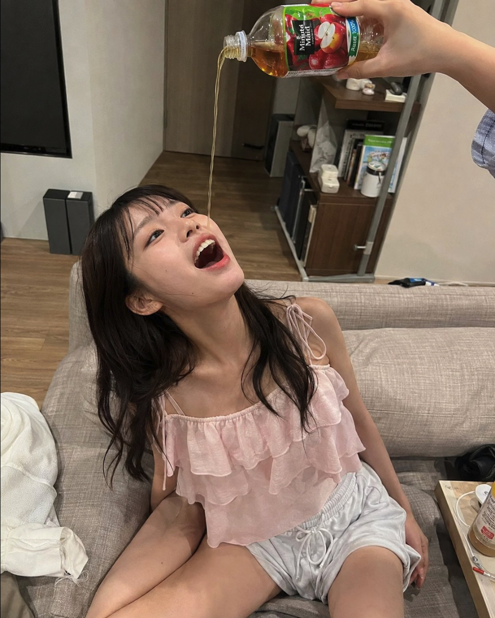 A woman with long dark hair sitting on a couch, mouth open, as apple juice is poured from above.