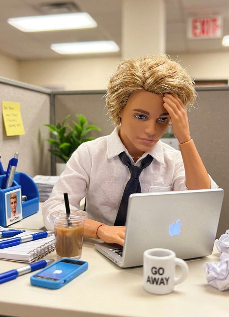 A blonde male fashion doll seated at a desk, resting his head against his hand in front of an open laptop.