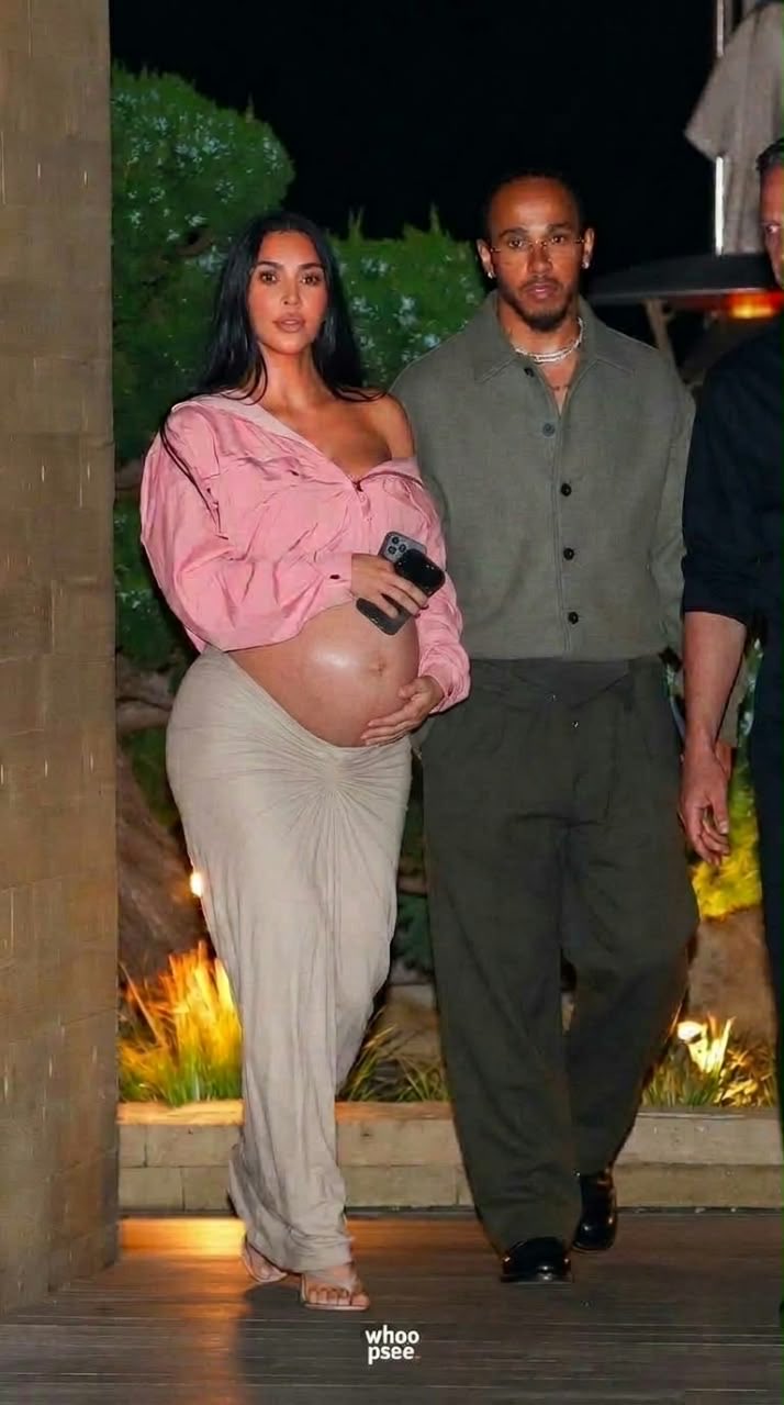 A pregnant woman resembling Kim Kardashian walking beside a man resembling Lewis Hamilton at night.