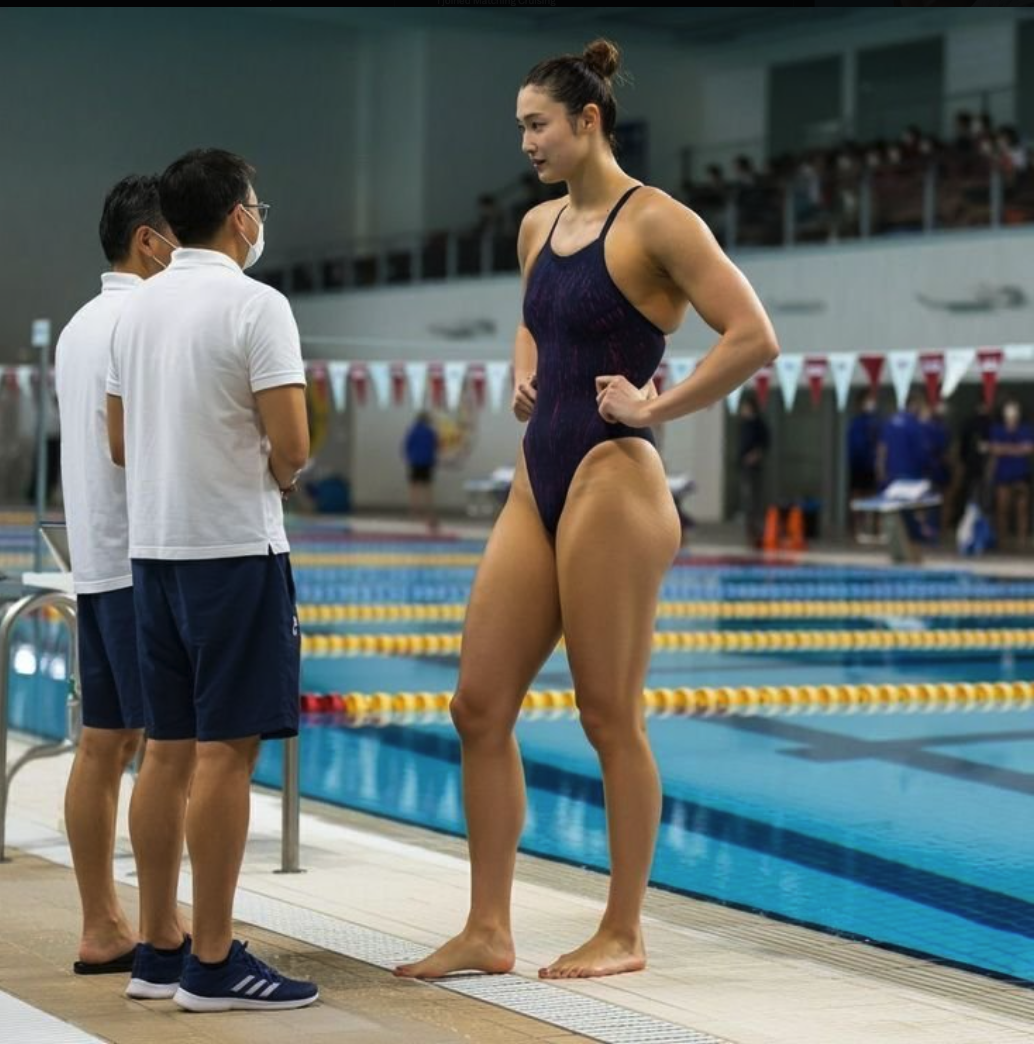 A woman in a dark swimsuit standing at the edge of a swimming pool.