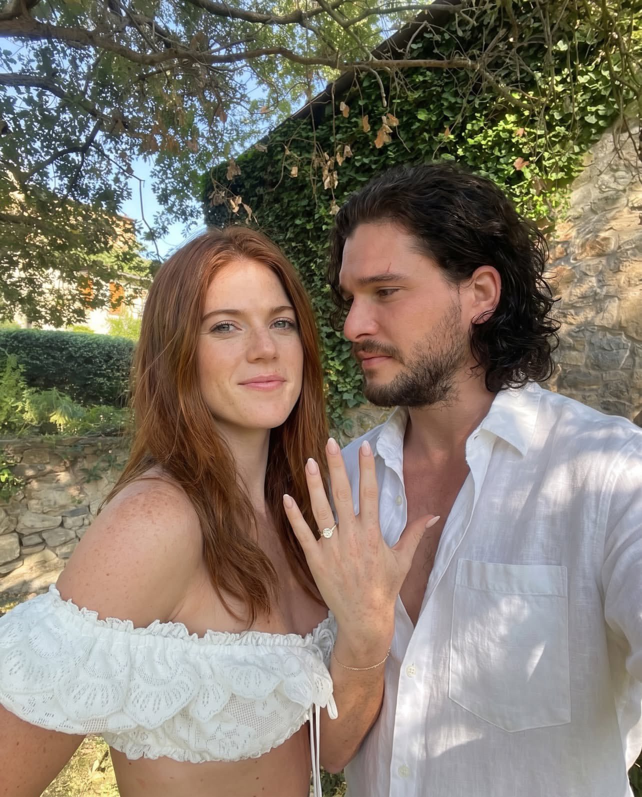 A woman resembling Rose Leslie and a man resembling Kit Harington posing closely together outdoors, with an engagement ring visible.