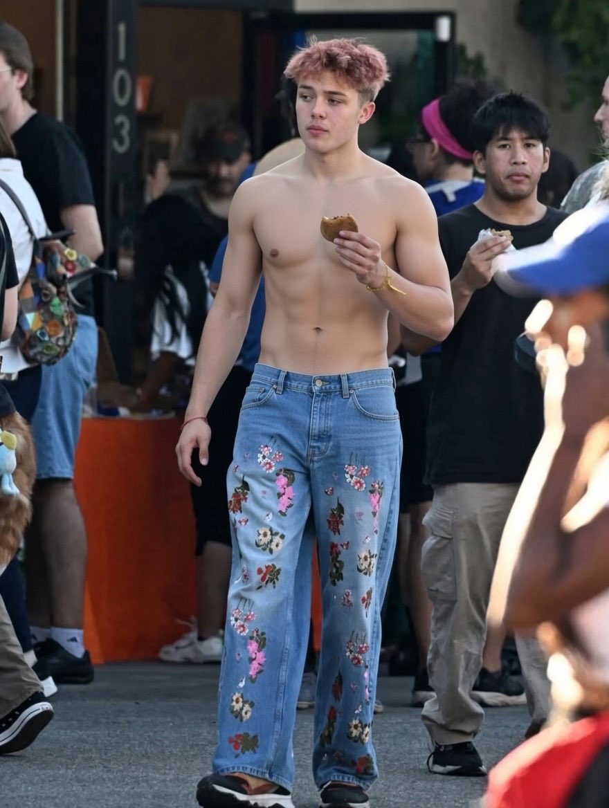 A shirtless man resembling Knox Léon Jolie-Pitt standing outdoors holding food.