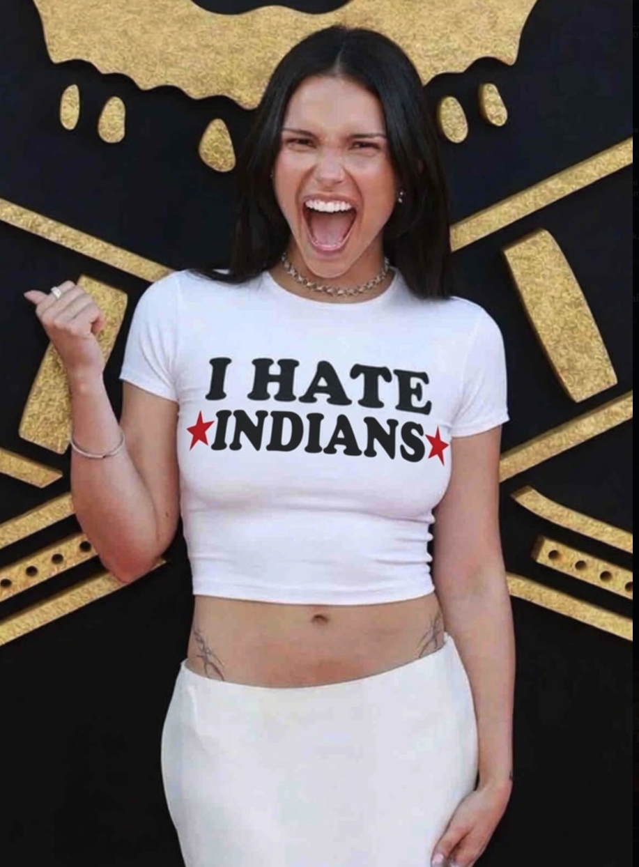 A woman resembling Kristina Anapau wearing a white crop top printed with 'I Hate Indians' against a black and gold background.