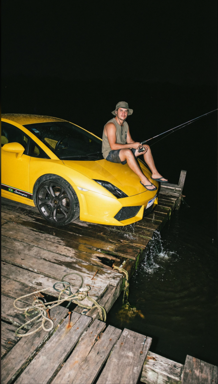 A man sitting on the hood of a yellow Lamborghini parked on a wooden dock, holding a fishing rod at night.