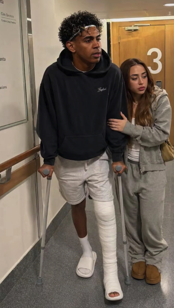 A young man resembling Lamine Yamal walking on crutches with a bandaged leg beside a young woman in a gray tracksuit.