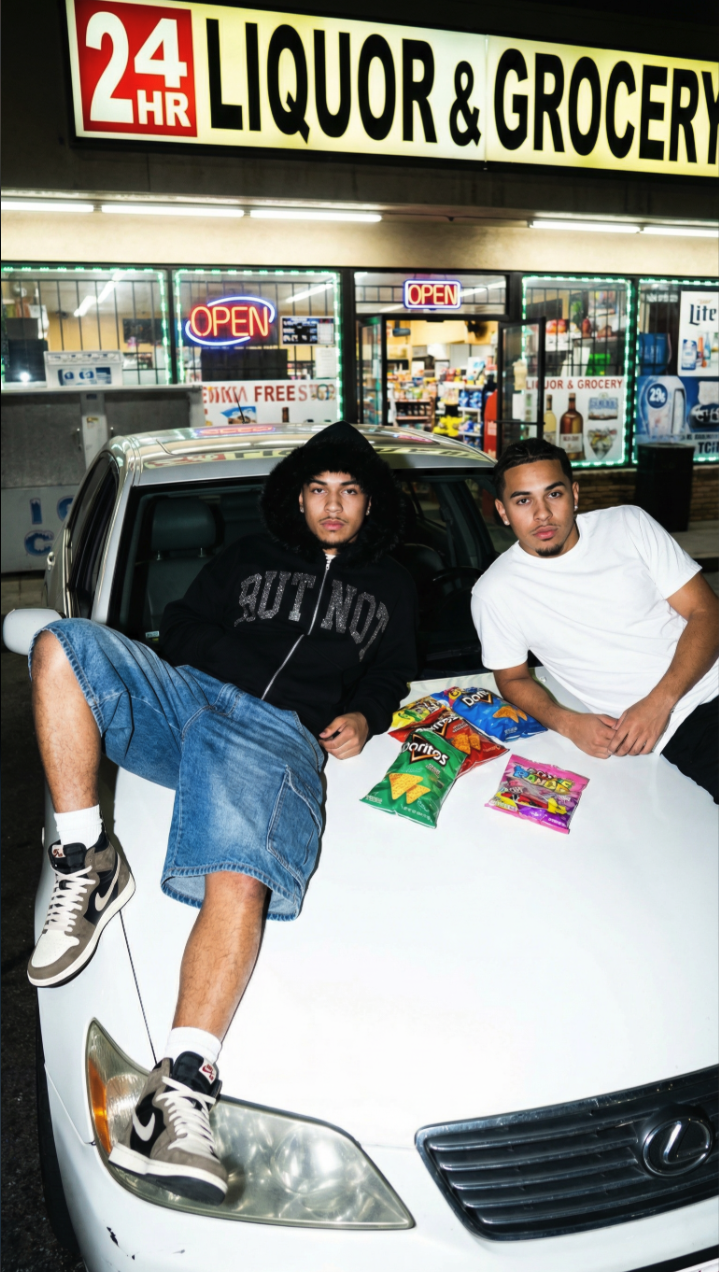 Two men, one resembling reggaeton singer Reykon, posing on a white car hood outside a 24-hour liquor and grocery store.