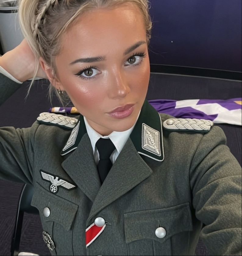 A young woman resembling Livvy Dunne in a military-style uniform with eagle and armband insignia, taking a selfie.