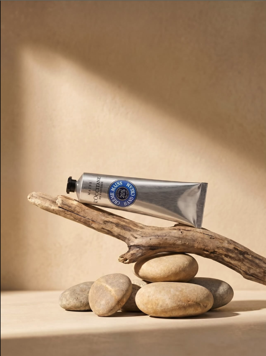 A silver tube of L'Occitane hand cream resting on a piece of driftwood balanced on smooth river stones.
