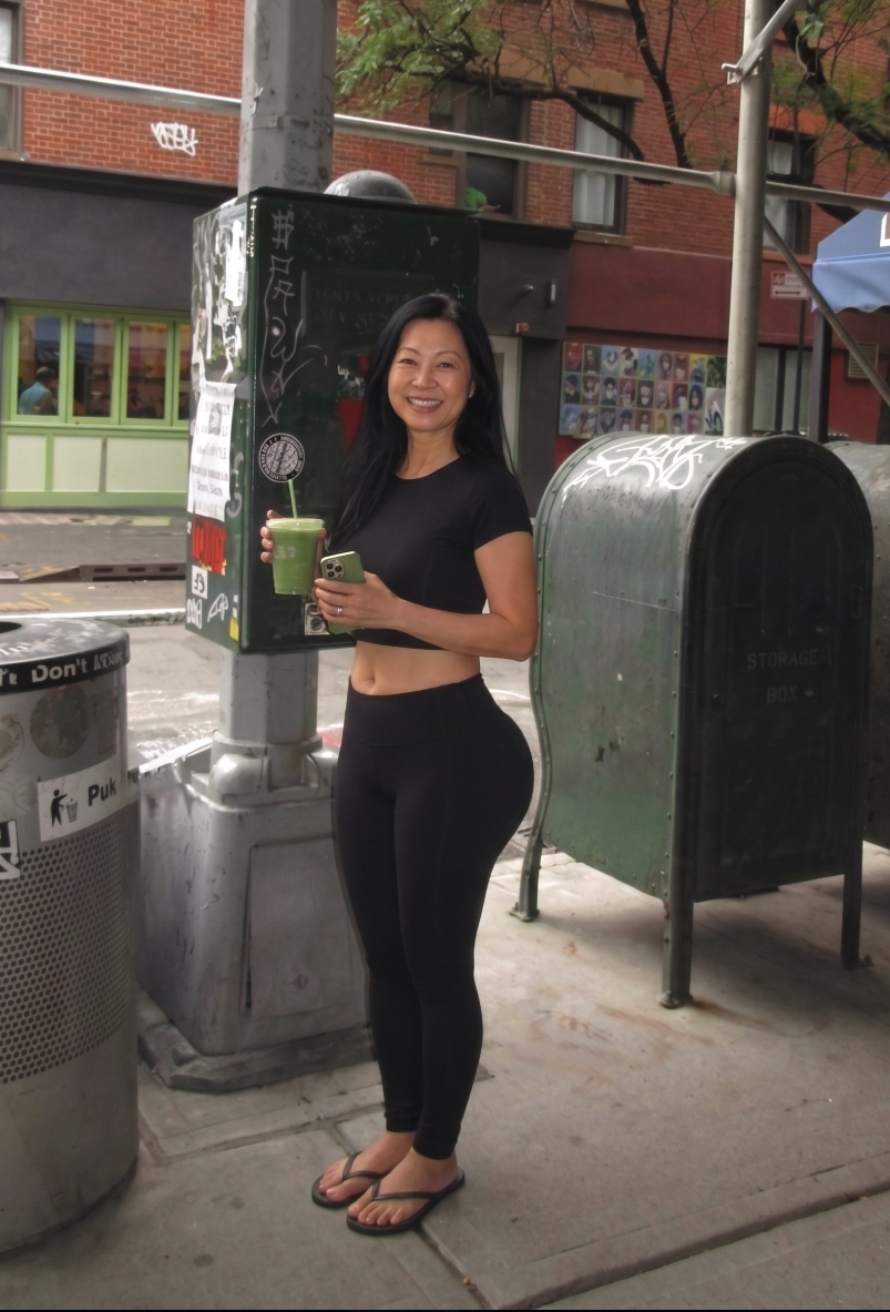 A woman standing on a city sidewalk holding a green drink and a phone.