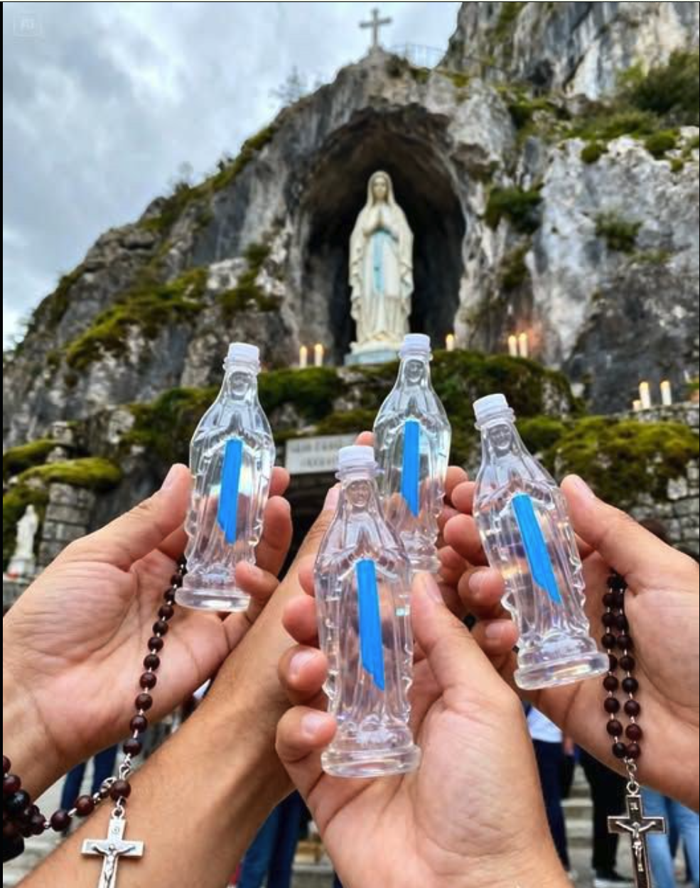 A statue resembling Our Lady of Lourdes in a rocky grotto, with hands in the foreground holding small Mary-shaped holy water bottles.