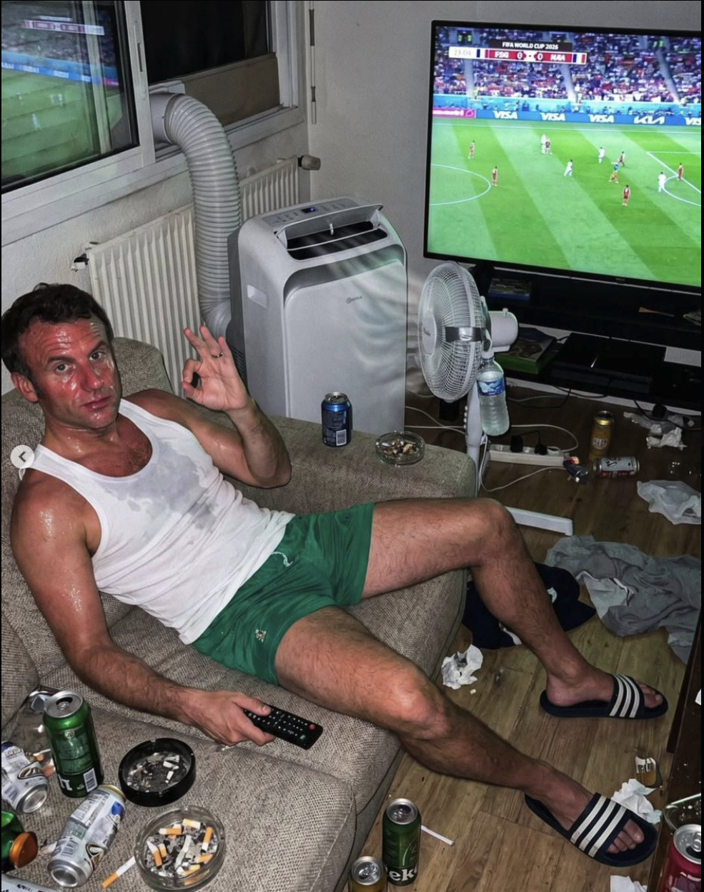 A man resembling Emmanuel Macron reclining on a couch in a cluttered living room, holding a TV remote and watching a soccer match.