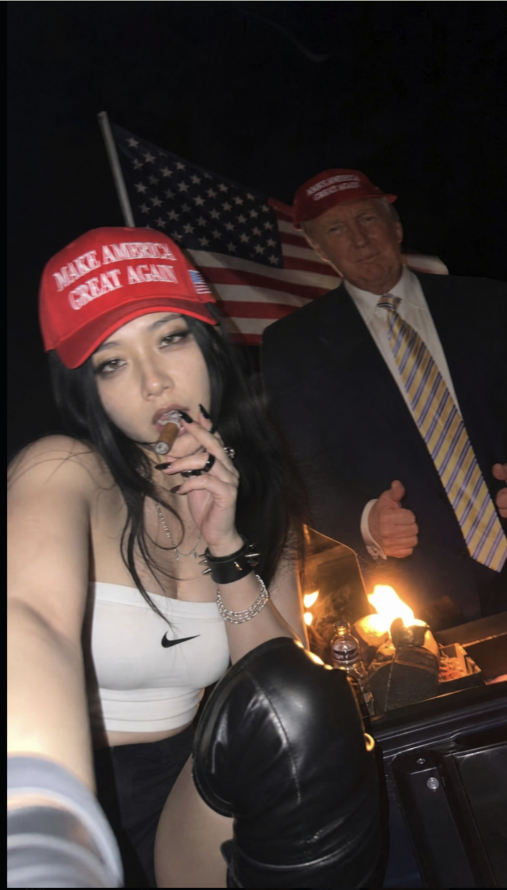 A young woman in a red 'Make America Great Again' cap taking a selfie, with a life-size cardboard cutout of a man in a suit and an American flag behind her.