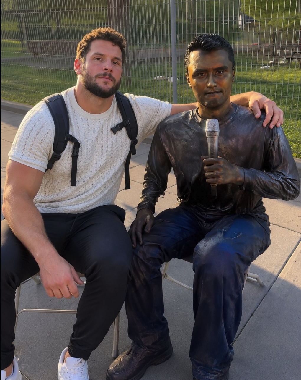 A man seated on a chair next to a bronze-colored statue of a figure holding a microphone, outdoors.
