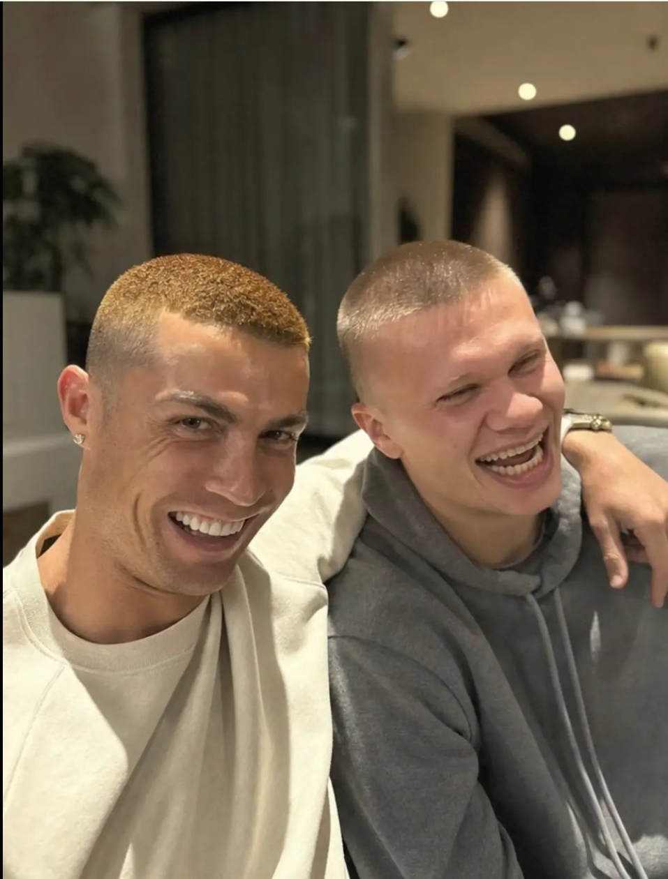 A man and a young person seated together indoors on a couch, both smiling and laughing toward the camera.