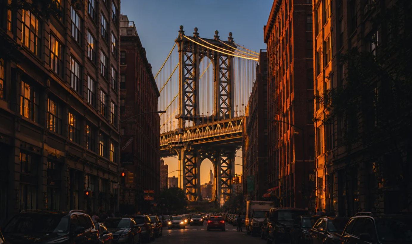 An AI-generated city street view looking toward the Manhattan Bridge tower at sunset, framed by brick buildings on both sides.