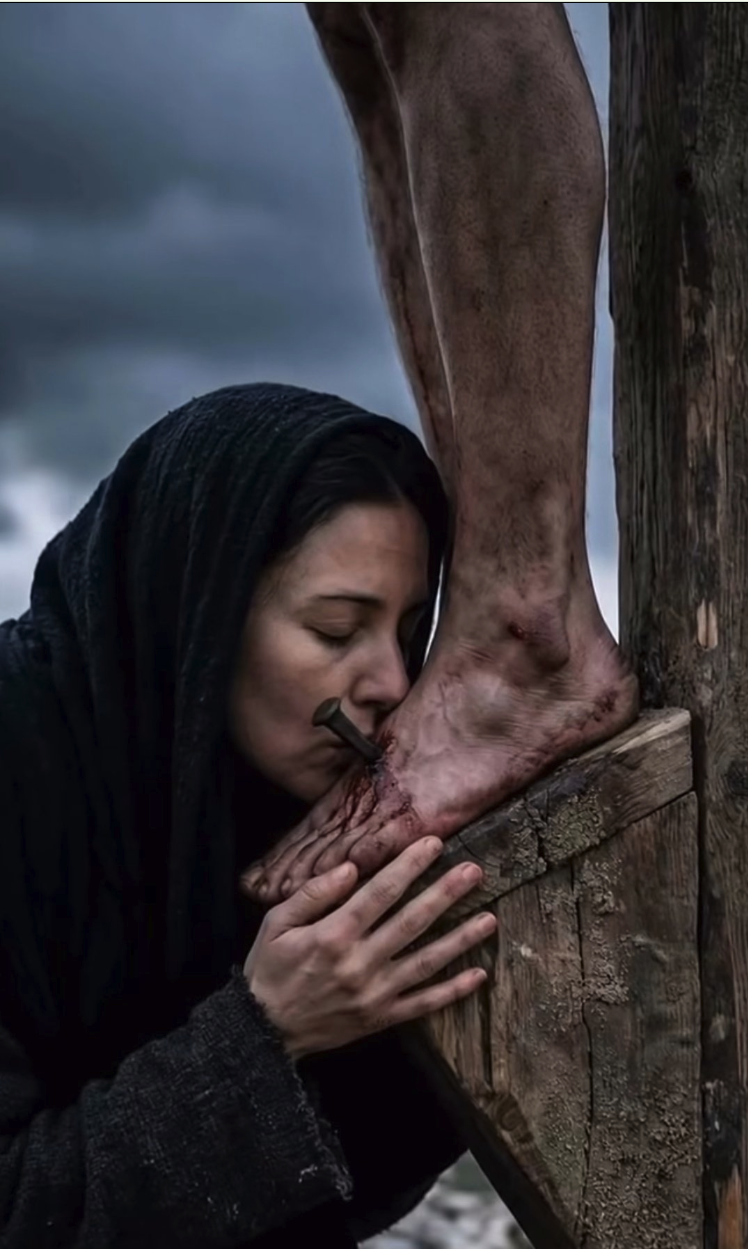 A woman resembling Marcella Michelangeli, in a dark hooded robe, kneeling and kissing the bleeding foot of a man nailed to a wooden cross.