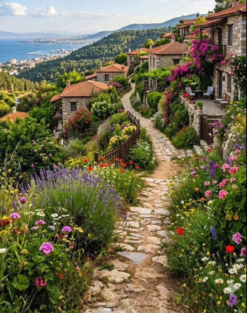 A cobblestone path winds through dense flower gardens past stone houses with terracotta roofs, overlooking a waterway and distant hills.
