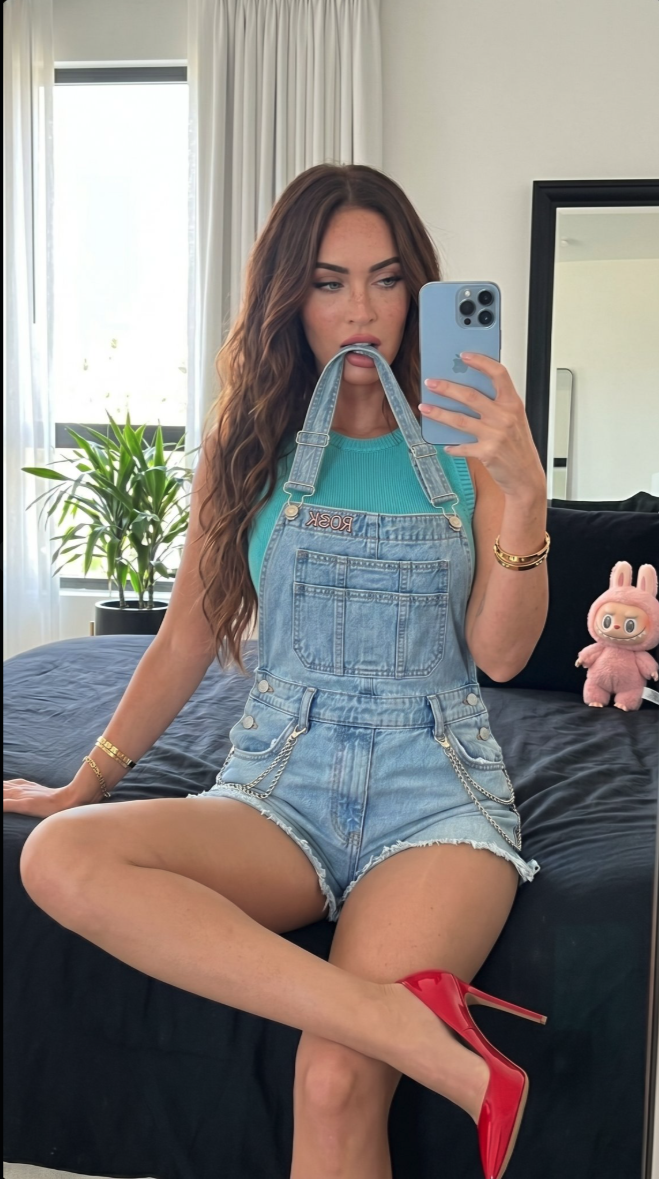 A woman resembling Megan Fox taking a mirror selfie on a bed, wearing denim overalls and a turquoise top with red high heels.
