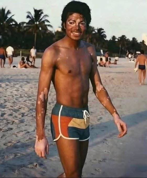 A shirtless man resembling Michael Jackson standing on a sandy beach in patterned shorts.