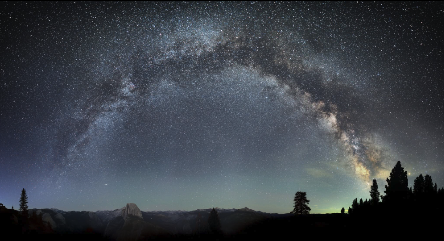 A panoramic view of the Milky Way arcing across a starry night sky above silhouetted mountains and evergreen trees.